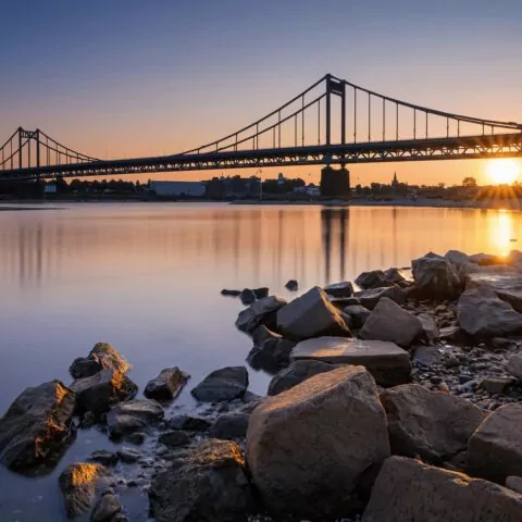 Bei Sonnenuntergang überspannt eine Hängebrücke einen ruhigen Fluss in Krefeld und wirft Spiegelbilder auf das Wasser. Der Himmel ist in warmen Orange- und Blautönen gehalten. Im Vordergrund säumen große Felsen das Ufer und verleihen der ruhigen Landschaft Struktur.