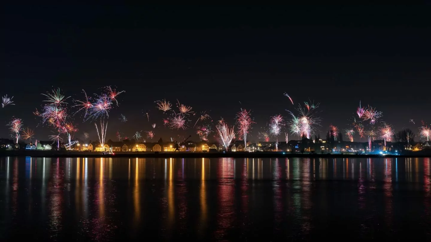 Ein Nachthimmel voller bunter Feuerwerke über der Stadt Leverkusen. Darunter erzeugen die Lichter der Gebäude entlang der Uferpromenade Spiegelungen im Wasser. Die Szene fängt die hellen Explosionen und die friedliche, beleuchtete Spiegelung auf der ruhigen Wasseroberfläche ein.