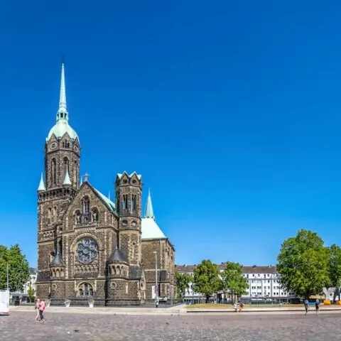 Ein Panoramablick auf den Stadtplatz von Mönchengladbach zeigt eine große historische Kirche mit einem hohen Turm und einem Uhrturm in der Mitte. Flankiert von anderen architektonischen Gebäuden und gesäumt von Bäumen bietet der Platz einen offenen, gepflasterten Raum unter einem klaren blauen Himmel.