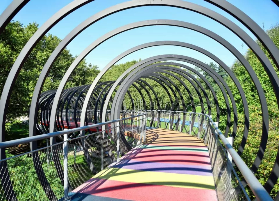 Eine Fußgängerbrücke in Oberhausen besteht aus einer Reihe von Metallbögen, die gemusterte Schatten auf einen regenbogenfarbenen Gehweg werfen. Der Weg ist von üppigen grünen Bäumen unter einem klaren blauen Himmel umgeben.