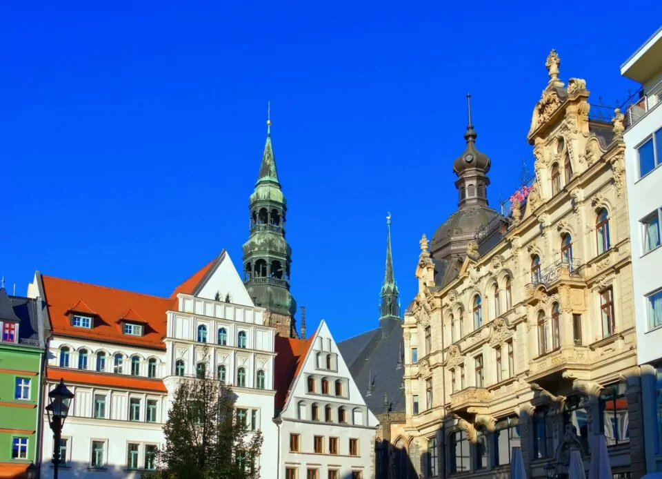 Eine malerische Stadtansicht von Zwickau mit historischen europäischen Gebäuden unter einem klaren blauen Himmel. Die Architektur weist unterschiedliche Stile auf, von kunstvollen klassischen Fassaden bis hin zu spitzen Dächern und Türmen, darunter ein markanter Uhrturm im mittleren Hintergrund.